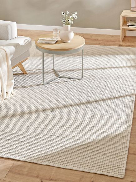 Runder Couchtisch mit Vase und Glas steht auf hellem, strukturiertem Teppich neben Sofa und Holzboden