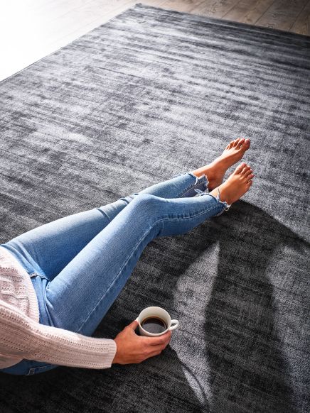 Person sitzt auf grauem Teppich und hält Kaffeetasse, trägt blaue Jeans und rosa Pullover.