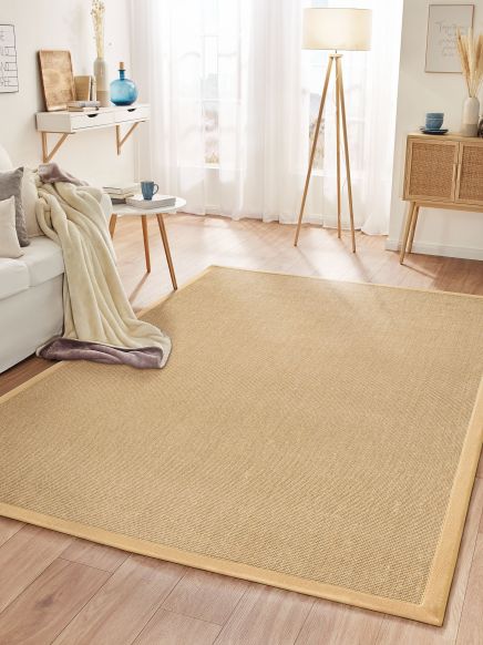 Beiger Teppich liegt auf Holzboden in hellem Wohnzimmer mit Sofa, Lampe und Kommode.