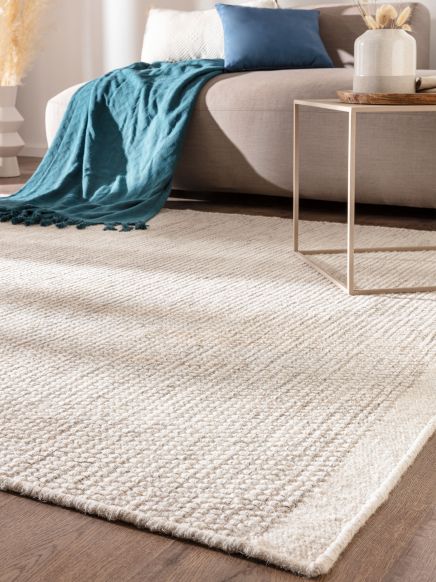 Beiger Teppich liegt auf Holzboden vor Sofa mit blauer Decke und Kissen, daneben Beistelltisch.
