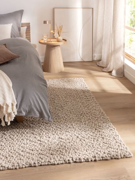 Teppich liegt auf Holzboden, neben Bett mit grauer Decke, Beistelltisch, Vorhang und Bild im Raum.