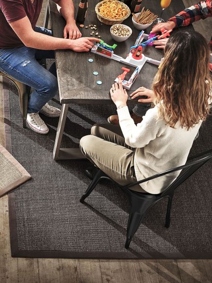 Drei Personen spielen ein Geschicklichkeitsspiel an einem Tisch mit Snacks und Getränken.