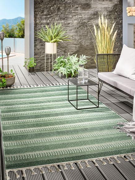 Grüner gestreifter Teppich liegt auf Holzterrasse mit Pflanzen, Sofa und kleinem Tisch.