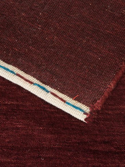 Rotbrauner Teppich zeigt umgeklappte Ecke mit weißem Rand und blauen sowie roten Fäden.