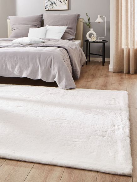 Bett mit grauer Bettwäsche, Nachttisch mit Lampe und Wecker, flauschiger Teppich, Holzfußboden.