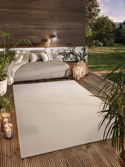 Terrassenmöbel liegen auf hellem Teppich, umgeben von Pflanzen und Laternen im Garten.