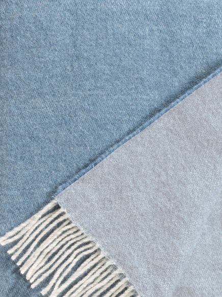 Zwei weich gewebte Decken in Blau und Hellgrau, eine mit Fransen, liegen übereinander.