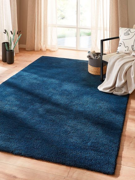 Blauer Teppich liegt auf Holzboden vor Fenster, daneben Stuhl mit Decke und Kissen.
