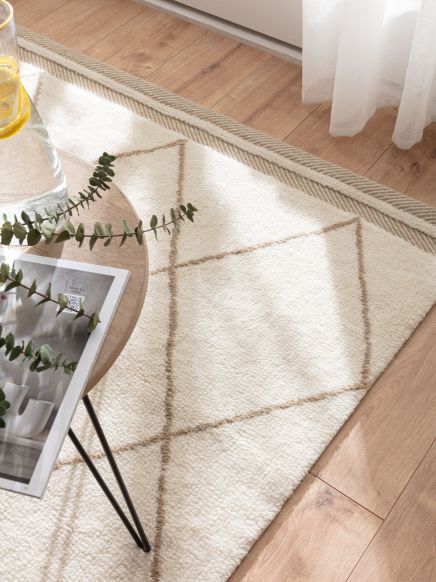 Tisch mit Glas und Pflanze steht auf cremefarbenem Teppich mit braunem Rautenmuster im Raum.