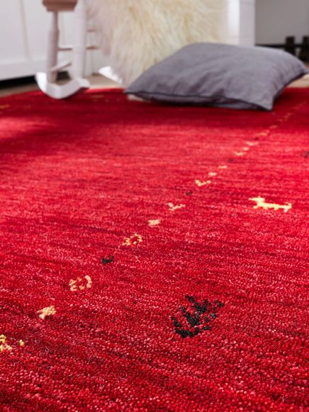 Roter Teppich liegt auf Holzfußboden, graues Kissen und weiße Schaukel mit Fell im Hintergrund.