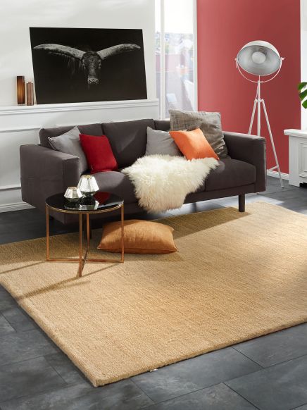 Dunkles Sofa mit Kissen und Fell liegt auf hellem Teppich, daneben kleiner runder Tisch mit Deko.