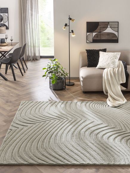 Beiger Teppich liegt auf Holzboden, Sofa mit Kissen und Decke, Pflanze und Stehlampe im Wohnzimmer.