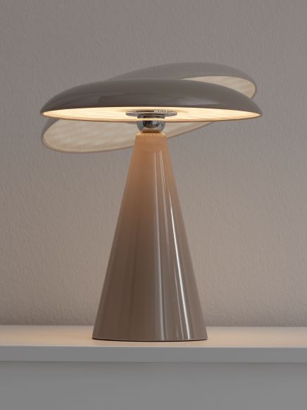 Lampe steht leuchtend auf weißem Möbel vor grauer Wand mit konischem Sockel und rundem Schirm.