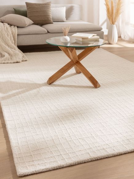 Runder Couchtisch mit Glasplatte steht auf hellem Teppich vor beige Sofa mit Kissen und Decke.