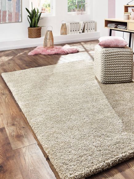 Heller Teppich liegt auf Holzboden, daneben Hocker mit Kissen und Schreibtisch im hellen Raum.