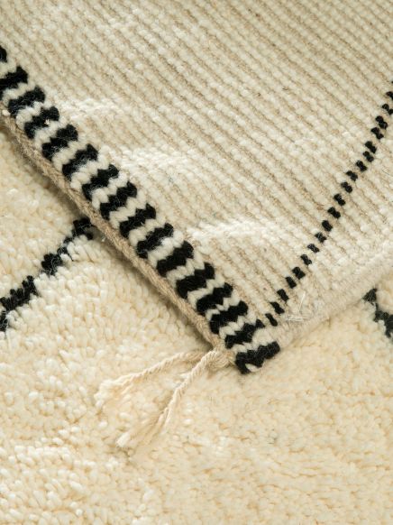 Zwei Teppiche liegen übereinander, beide cremefarben mit schwarzen Mustern und Fransen.