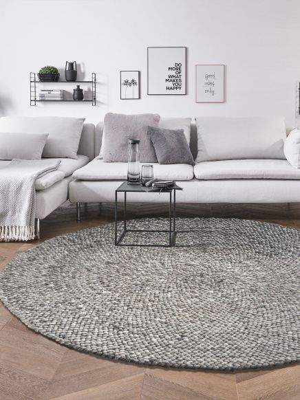 Runder grauer Teppich liegt auf Holzboden vor grauem Sofa mit Kissen und kleinem schwarzen Tisch.