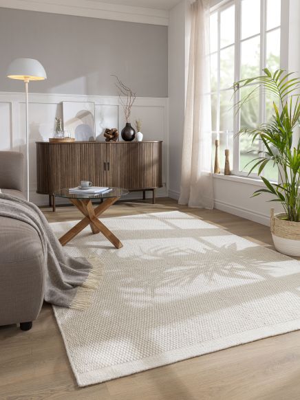 Beistelltisch trägt Tasse im Wohnzimmer mit Sofa, Teppich, Sideboard, großer Pflanze und Fenster.