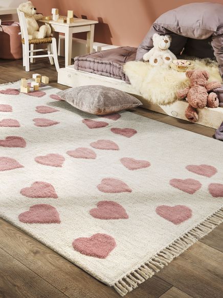 Teppich mit rosa Herzmustern liegt auf Holzboden, umgeben von Kissen, Kuscheltieren und Spielzeugwürfeln.