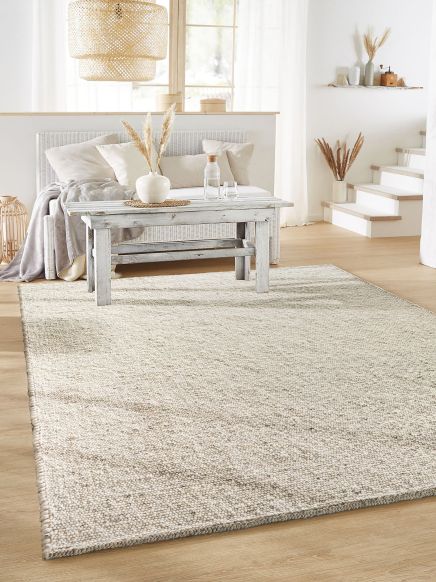 Heller Teppich liegt vor einem Sofa mit Kissen und Decke, Holztisch mit Vase im Raum.