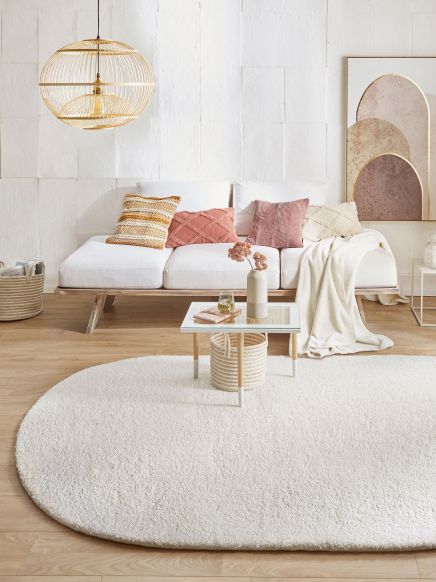 Rundes cremefarbenes Teppich liegt vor weißem Sofa mit bunten Kissen in hellem Wohnzimmer.