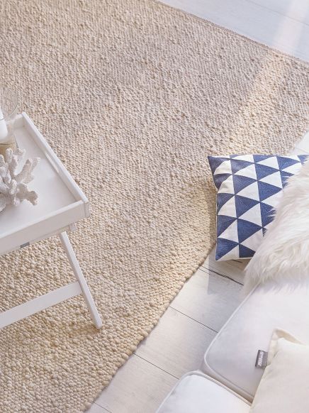 Beiger Teppich liegt auf hellem Holzboden, neben Sofa mit Kissen und weißem Tisch mit Deko.