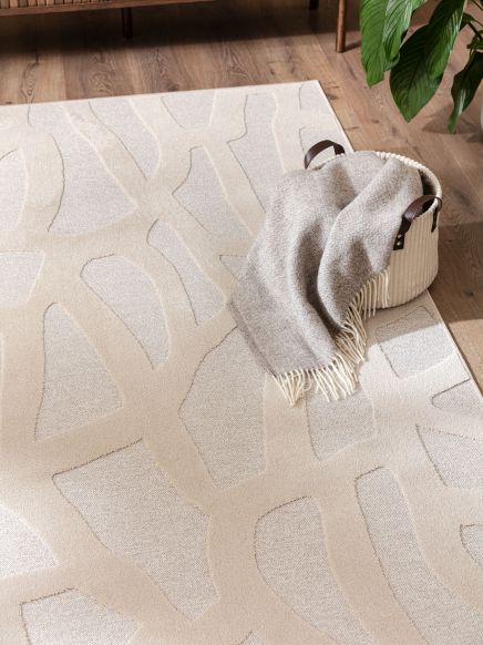 Korb liegt auf beige Teppich mit abstraktem Muster, gefüllt mit grauer Decke, Holzfußboden und Pflanze.