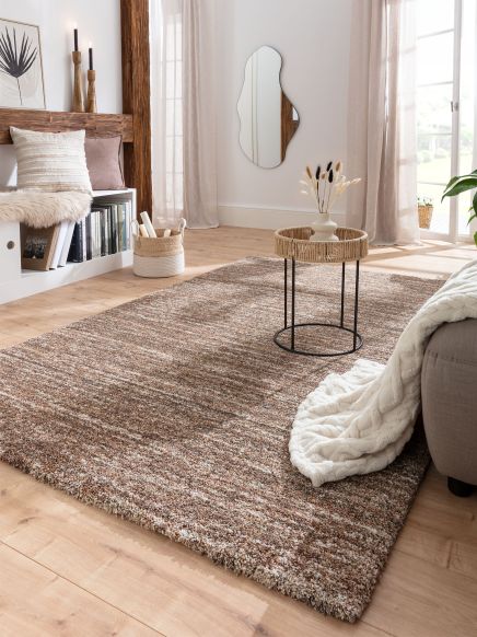 Braun gemusterter Teppich liegt auf Holzboden, kleiner Tisch mit Vase und getrockneten Blumen in hellem Wohnzimmer.