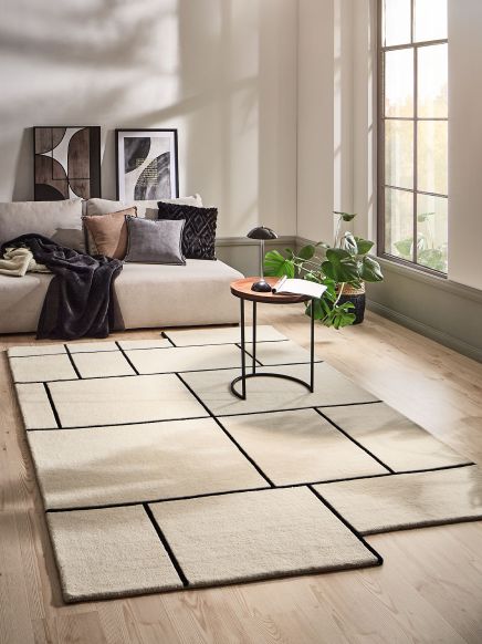 Beiger Teppich mit schwarzem Linienmuster liegt vor Sofa mit Kissen, daneben kleiner Tisch und Pflanze.