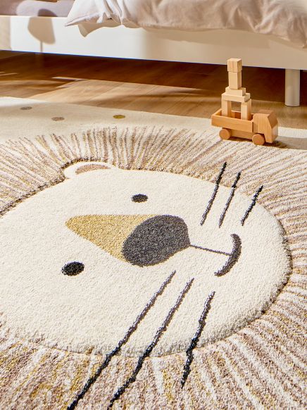Kinderteppich mit Löwengesicht liegt auf Holzboden, daneben Holzspielzeug neben Bett.