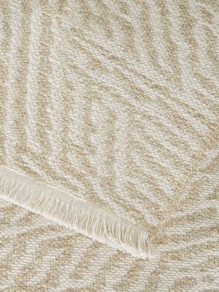 Teppich legt sich über weiteren Teppich mit beige-weißem Fischgrätmuster und Fransenrand.