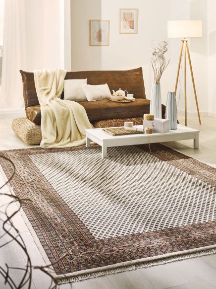 Braunes Sofa steht mit Decke und Kissen neben weißem niedrigen Tisch auf gemustertem Teppich im hellen Raum.