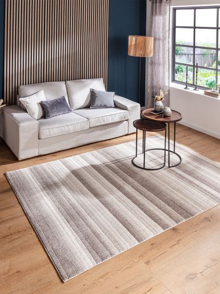 Beiger Sofa sitzt vor Holzpaneel, zwei runde Tische mit Deko stehen auf gestreiftem Teppich im Raum.