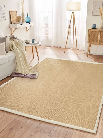 Beiger Teppich liegt auf Holzboden, daneben Sofa mit Decke, Tisch, Lampe und Sideboard im hellen Wohnzimmer.
