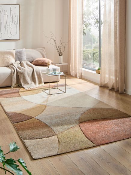 Teppich liegt auf Holzboden, Sofa mit Decke und Kissen, kleine Tische mit Vase, große Fenster mit Vorhängen.
