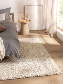 Teppich liegt auf Holzfußboden neben Bett und Beistelltisch mit Glas, Lampe und Vase im hellen Zimmer.