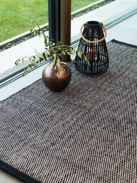 Vase mit Zweig steht neben Laterne mit Kerze auf Teppich vor bodentiefem Fenster.