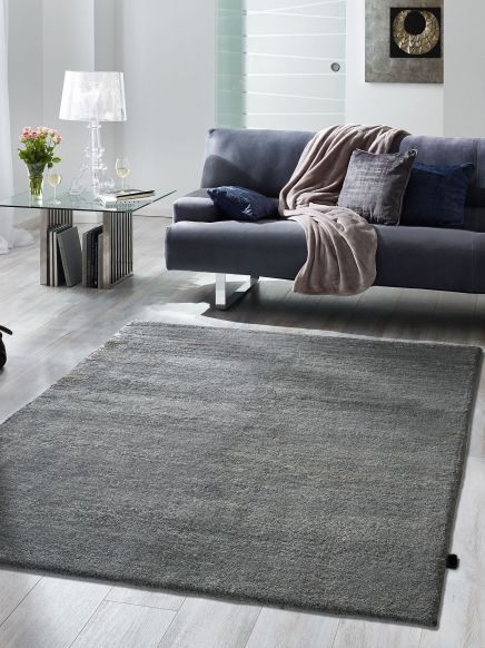 Grauer Teppich liegt auf hellem Boden vor einem grauen Sofa mit Decke im modernen Wohnzimmer.