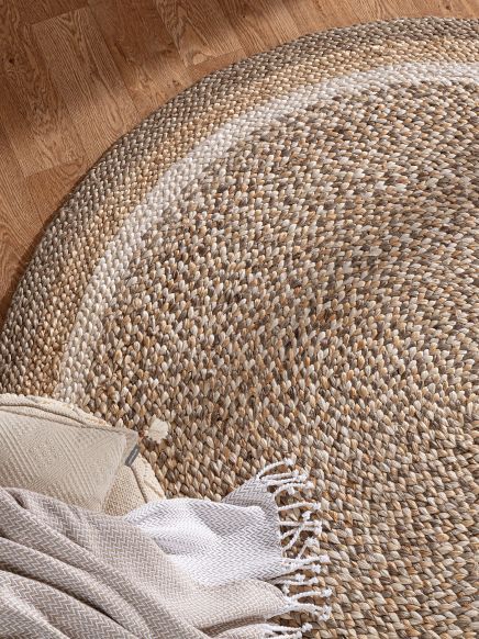 Runder Teppich liegt auf Holzboden, daneben beige Decke und Kissen mit Muster.