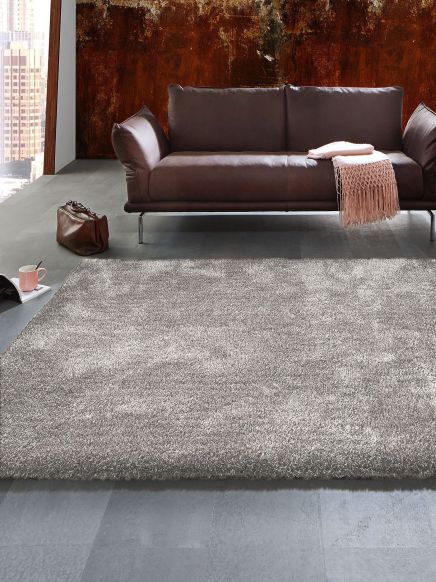 Braunes Sofa steht mit Decke und Tasche auf grauem Teppich in modernem Raum mit Stadtblick.
