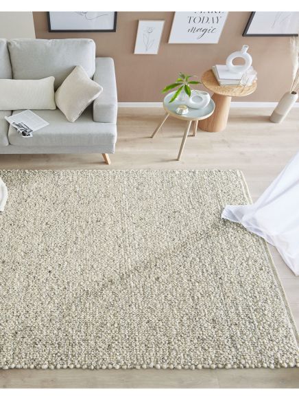 Hellgrauer Teppich liegt auf hellem Holzfußboden, neben grauem Sofa mit Kissen und zwei Tischen.