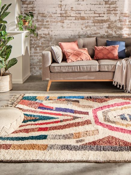 Wohnzimmersofa mit Kissen und Decke vor buntem Teppich, Pflanzen und Backsteinwand im Raum.