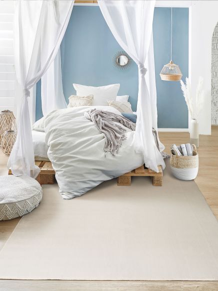 Bett mit weißer Bettwäsche und Vorhängen steht auf Holzpalette vor blauer Wand in hellem Zimmer.