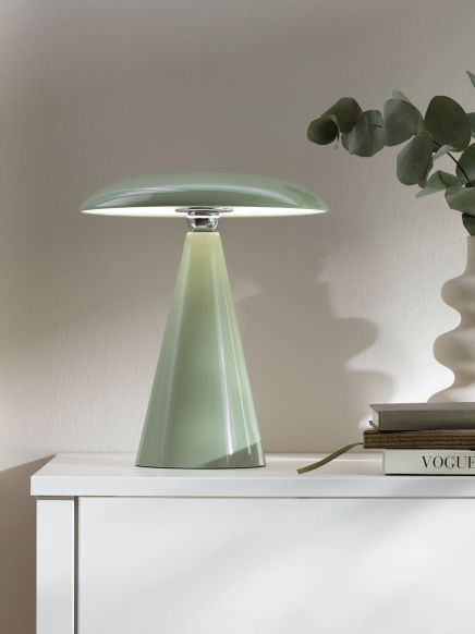 Moderne grüne Tischlampe leuchtet auf weißem Sideboard neben Büchern und Vase mit Blättern.