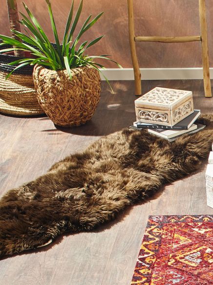 Flauschiger Teppich liegt auf Holzboden, daneben Pflanze in Korb, Bücher und Box, Bereich im Raum.