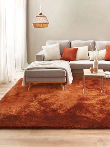 Grau-beiges Ecksofa mit Kissen liegt auf rostfarbenem Teppich neben kleinem Beistelltisch im Zimmer.