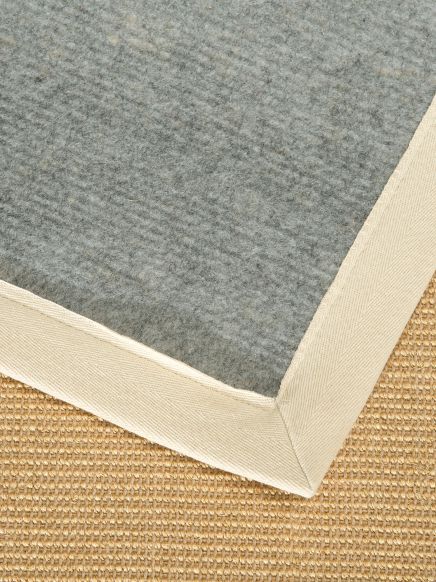 Ecke eines grauen Teppichs mit hellbeiger Umrandung auf einem hellbraunen Bodenlaminat.