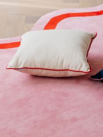 Kissen mit roter Kante liegt auf rosa Teppich mit rotem Streifen, Holzboden im Hintergrund sichtbar.