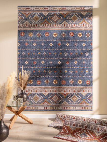 Blau gemusterter Teppich hängt an Wand, daneben Glastisch mit Vasen und Pampasgras, helle Wand.