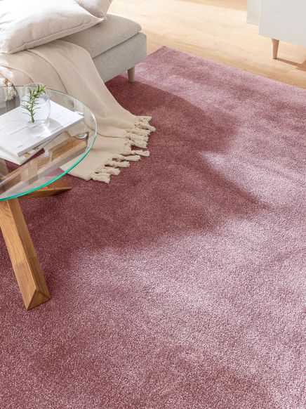 Rosa Teppich liegt unter grauem Sofa mit beiger Decke und Glas-Couchtisch mit Vase.